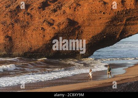 Couple in love walking under huge arch on Legzira ocean beach at sunset. Famous and popular landmark in Morocco. Stock Photo