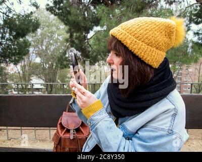 Young adult woman with yellow woolen cap sitting on a park bench looking into the mirror. Autumn urban concept. Stock Photo