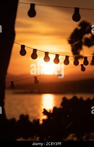 Silhouette of vintage light bulbs on string wire and pine trees against sunset over the sea in background. Vacation, tourism, travel and summer Stock Photo