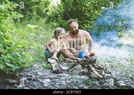 Father and little cute son sitting by the fire in the forest. Best friends spending time together. Stock Photo