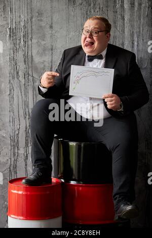 businessman happy with increasing of oil prices on global market, handsome guy sits on oil barrel, holding analysis chart showing current position Stock Photo