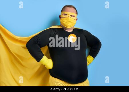 handsome brave strong man has a mad desire, a dream to shield the world from dirt and evil. fat superhero in yellow costume posing isolated over blue Stock Photo