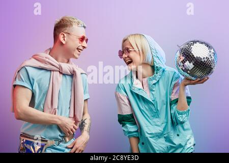A couple of young stylish blonde female and albino male dancers wearing pink sunglasses and mint blue and pink clothes poses with disco ball on purple Stock Photo