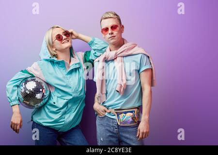 A couple of young stylish blonde female and albino male dancers wearing pink sunglasses and mint blue and pink clothes poses with disco ball on purple Stock Photo