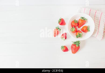 Top view of fresh strawberries on a white rustic wooden table, inside a bowl ready to eat as organic and healthy food, next to a cloth with red stripe Stock Photo