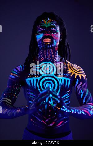 portrait of man having fluorescent make-up and body art glowing on neon lights, african guy stand in the pose of prayer Stock Photo