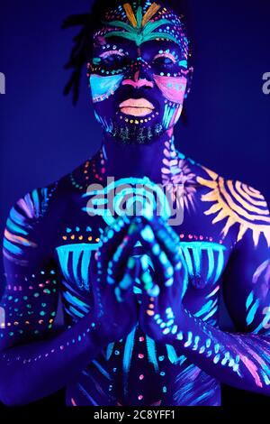 portrait of man having fluorescent make-up and body art glowing on neon lights, african guy stand in the pose of prayer Stock Photo