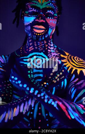 portrait of young african man in fluorescent body art with crossed hands, look at camera. strong male in darkness, neon lights Stock Photo