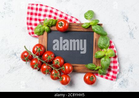 Ripe cherry tomatoes and fresh basil leaves on stone table with chalkboard, cooking ingredients top view