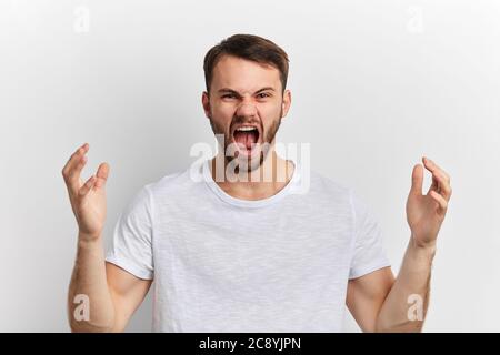 Young man wearing white T-shirt raising his arms, shouting, expressing aggressive expression standing over white isolated background. Frustration conc Stock Photo