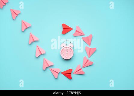Flat lay with pink and red paper airplanes around a pink clock on a blue background. Time flies concept with paper planes flying around the alarm cloc Stock Photo