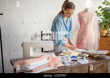 Pleasantly looking wearing ponytail is sitting at her worktable with laid tailoring equipment, containers full of thread spools, thinking of color whi Stock Photo