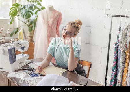 Pensive beautiful blonde dressmaker in fashion atelier in stylish eyeglasses and mint casual t-shirt drawing sketch of wedding dress Stock Photo