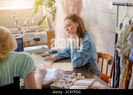 Cute female seamstress discusses with the client sketch of custom made lady s suit. Young female tailor and cloth designer collaborating in the worksh Stock Photo