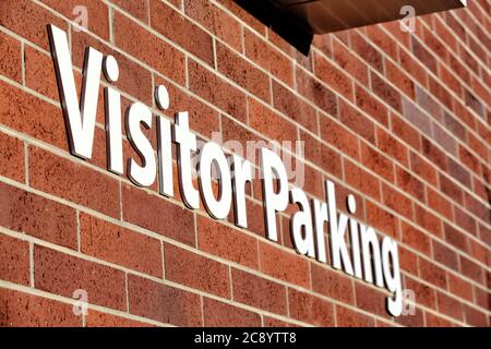 A white visitor parking sign in block letters, on a red brick wall, directs visitors to a reserved parking area.