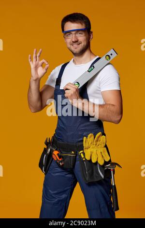 Young caucasian repairman worker in safety glasses and uniform holding builder level. handyman wearing tool belt showing Ok gesture isolated on orange studio background Stock Photo