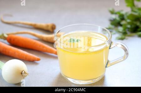 Clear homemade bone  broth in the glass cup on the table. Carrot,onion and parsley are around. Horizontal orientation. Stock Photo