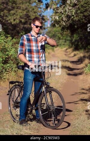 The young guy sits by the standing bicycle on a track in the wood among trees and looks at time on a watch. Stock Photo