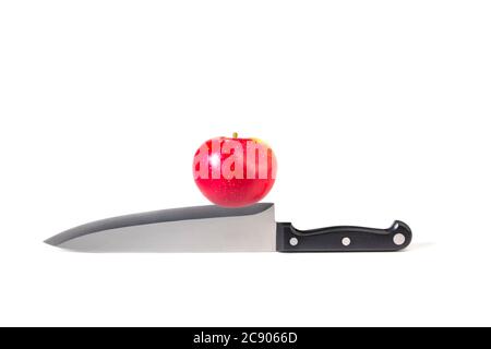 Red apple resting on cutting blade edge of large kitchen knife on white background. Concept photography: at the cutting edge, on a knife-edge. Stock Photo