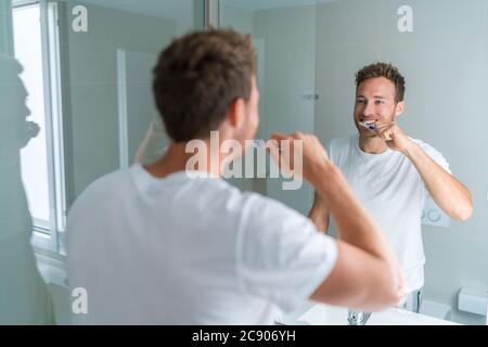 Man getting ready in the morning doing hygiene routine brushing his teeth looking in mirror of home bathroom using toothbrush in for clean dental oral Stock Photo
