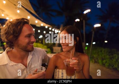 Couple dating drinking at bar on night out at outdoor restaurant terrace in Hawaii vacation travel. Asian woman, man having fun together toasting mai Stock Photo