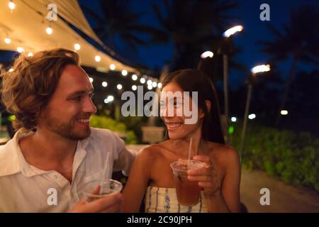 Couple dating drinking at bar on night out at outdoor restaurant terrace in Hawaii vacation travel. Asian woman, man having fun together toasting mai Stock Photo