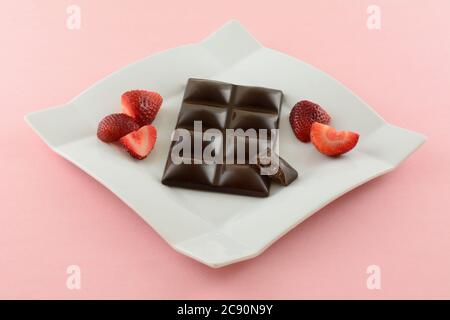 Dark chocolate candy bar with strawberry filling and fresh raw strawberries on white dessert plate on pink background Stock Photo