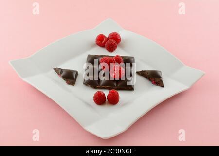 Dark chocolate candy bar with bits of raspberry and fresh raw red raspberries on white dessert plate on pink background Stock Photo