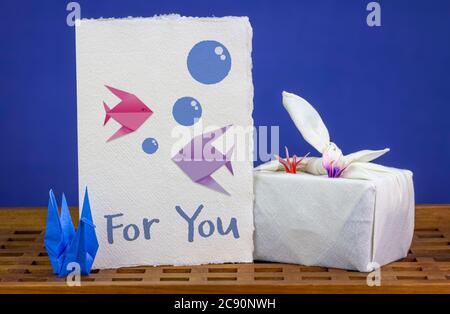 Handmade origami swans, celebration card with origami fish together with Furoshiki fabric wrapped gift, on wooden tray. Reuse textiles and paper for h Stock Photo