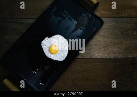 Fried egg white and yolk on rectangular cast iron flat griddle pan with wooden handles, wood tabletop background, flat lay. Minimalism, minimalist Stock Photo