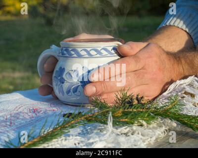 Close up hand of male holding hot smoking tea in rustic white blue painted ceramic cup, white tablecloth, green pine branches, cone and cloves spices Stock Photo