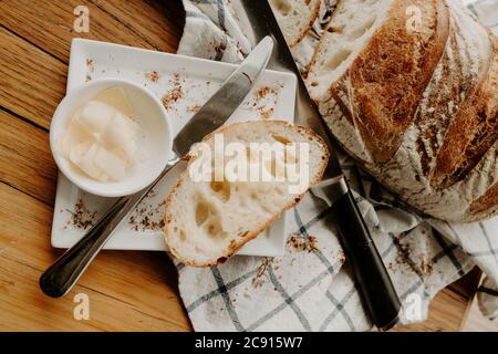 A slice of bread on a white plate with butter with Home made Sour dough bread cooling on a tea towel which is on a wooden chopping platter board, wild Stock Photo