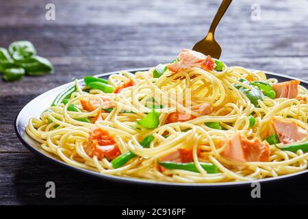 Salmon pasta of spaghetti, green beans, roasted fish and garlic with fresh basil on top served on a black plate on a dark wooden table, close-up Stock Photo
