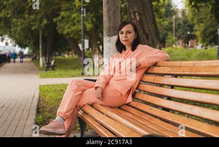 Young woman sits on bench in park. Adult female enjoys her rest on fresh air. Stock Photo
