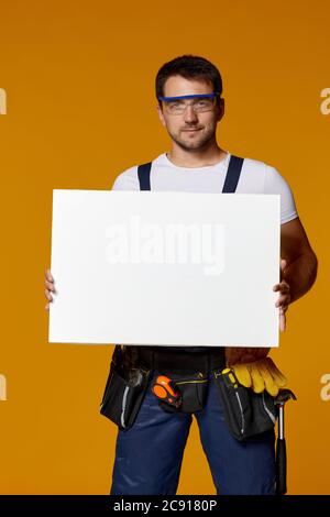 caucasian repairman worker in uniform and safety glasses holding empty blank board. handyman wearing tool belt isolated on yellow studio background Stock Photo