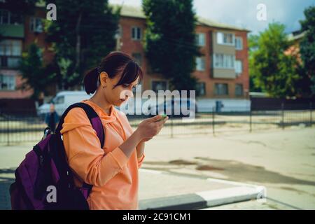Attractive tourist with a backpack on a city street with a mobile phone in her hands. Side view of a woman focusing on a smartphone screen while Stock Photo