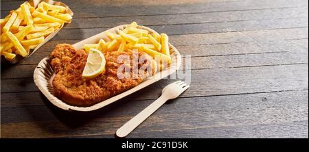 Serving of crumbed fried fish or schnitzel with French fries and a small plastic fork on a cardboard disposable plate over a rustic wooden table with Stock Photo