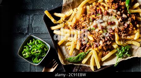 Close up on savory mince, cheese and potato fries seasoned with hot chili peppers and herbs served on paper in an oven tray in panorama banner format Stock Photo