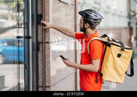 Modern food delivery. Young man with beard, with backpack and smartphone, ringing the bell Stock Photo