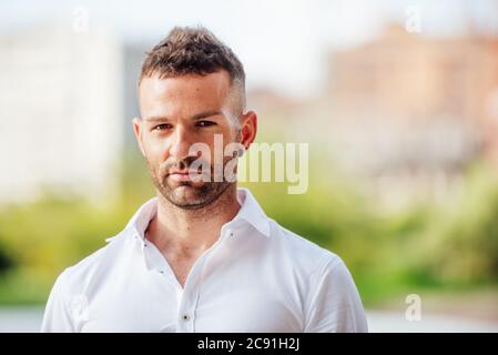 young man in white shirt looking at camera with short hair and short beard outdoors with plants and flowers background Stock Photo