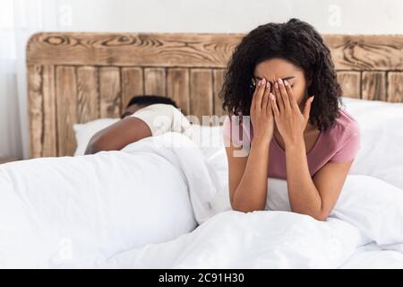 Crying african woman sitting on bed next to sleeping man