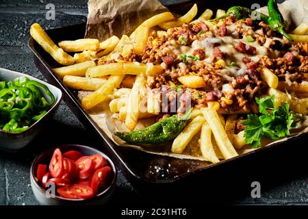 Serving of crispy French fries topped with minced beef, chili peppers and cheese in close up with sides of fresh peppers Stock Photo
