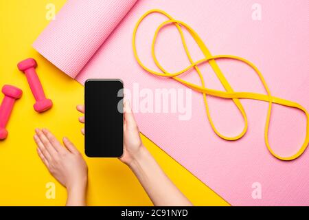 cropped view of woman holding smartphone near resistance band on pink fitness mat and dumbbells on yellow background Stock Photo