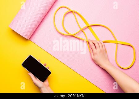 cropped view of woman holding smartphone and resistance band on pink fitness mat on yellow background Stock Photo