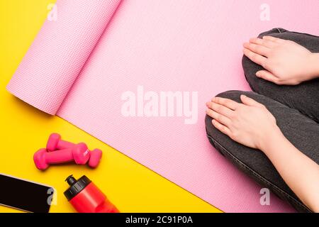 partial view of woman sitting on pink fitness mat near sports bottle, smartphone, dumbbells on yellow background Stock Photo