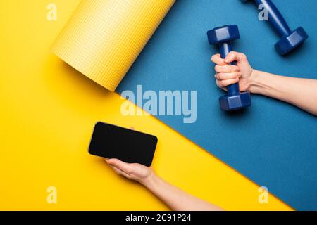 cropped view of woman holding dumbbell and smartphone on blue fitness mat on yellow background Stock Photo