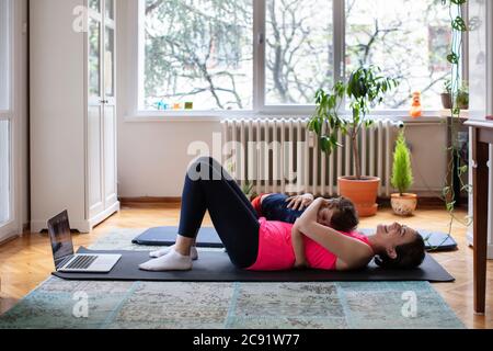 Happy Mother and Child Cuddling with Joy while Exercising Pilates, Yoga, Fitness at Home in front of the Laptop Stock Photo