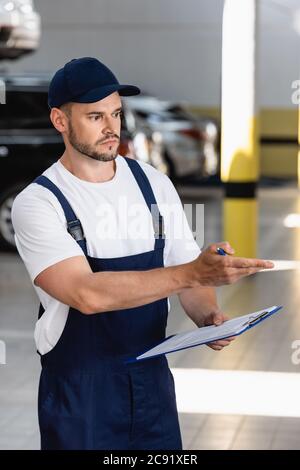 handsome mechanic in uniform and cap pointing with hand while holding clipboard and pen Stock Photo