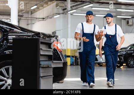 selective focus of handsome mechanics in uniform using digital tablet and walking near cars in workshop Stock Photo