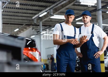 selective focus of handsome mechanics in uniform using digital tablet and looking at car in workshop Stock Photo